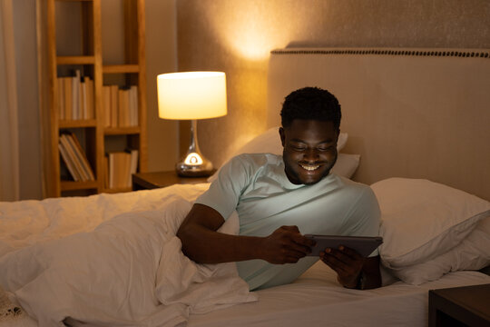African American man reclining in bed, holding tablet and smiling under warm bedside lamp