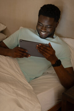 African American man reclining on bed wearing light mint T-shirt, holding tablet, smiling