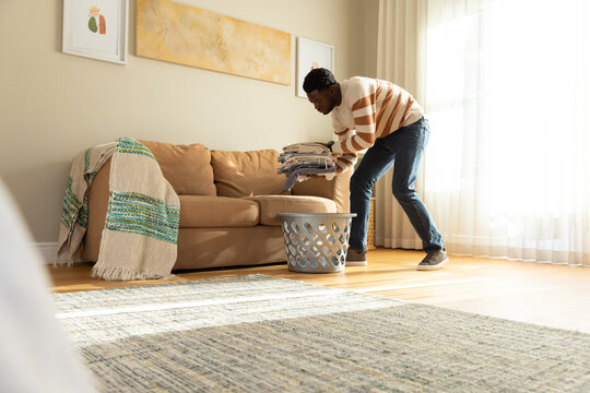 African American man folding clothes in living room beside sofa and gray laundry basket, copy space