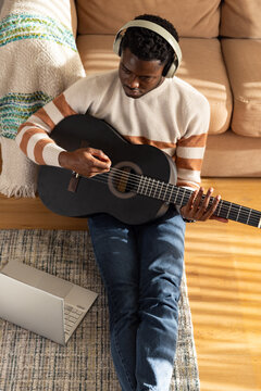 Adult African man playing guitar on living room floor wearing headphones, sweater near open laptop