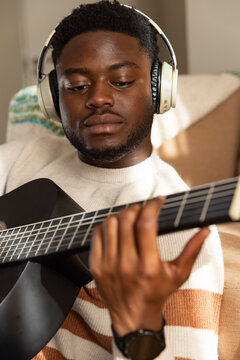 Male sitting on couch at home wearing striped knit sweater and headphones, fingering black guitar