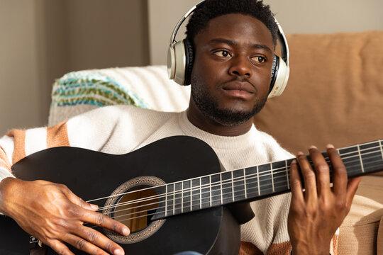 African American man playing black acoustic guitar on sofa wearing headphones and cream sweater