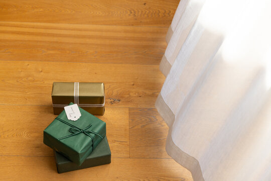 Two green boxes and gold box are resting on hardwood floor near sheer curtain with ribbon