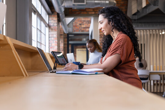 African American women colleagues working at desk in rust top with laptop and tablet, copy space