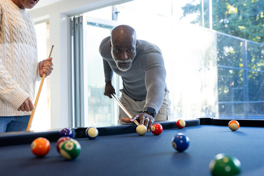 Diverse male friends playing pool in home game room on dark-blue table, lining shot with cues