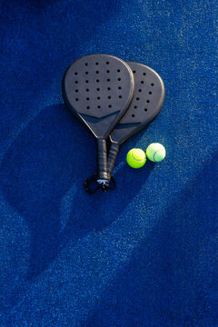 Two black padel rackets are lying overlapping on blue textured court, two yellow balls nearby