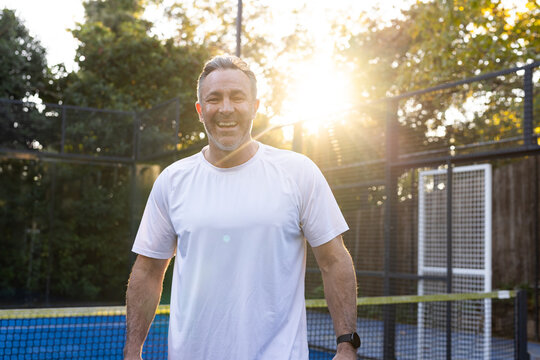 Middle-aged man standing wearing white T-shirt smiling on blue court near net, wristwatch