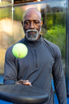 Senior African man tossing neon ball holding black paddle on blue court by glass, wearing gray-top