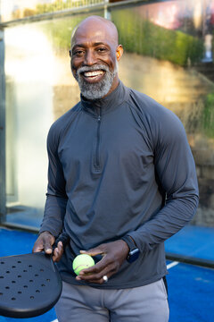 African American senior male smiling, holding black paddle, yellow ball on blue court in gray top