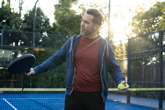 Adult male holding paddle, yellow ball on blue court, getting ready to serve in navy hoodie