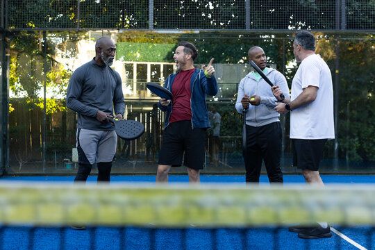 Group of four men standing, holding padel rackets, ball and net, wearing sportswear on blue court