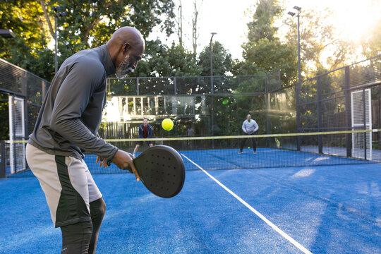 Diverse men playing on padel court, left man preparing with racket and yellow ball, copy space