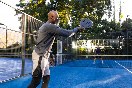 Diverse male opponents competing on blue padel court, hitting yellow ball with perforated paddle