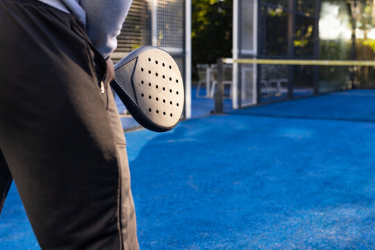 African male holding padel racket at hip on blue court near net, gray sweatshirt, copy space