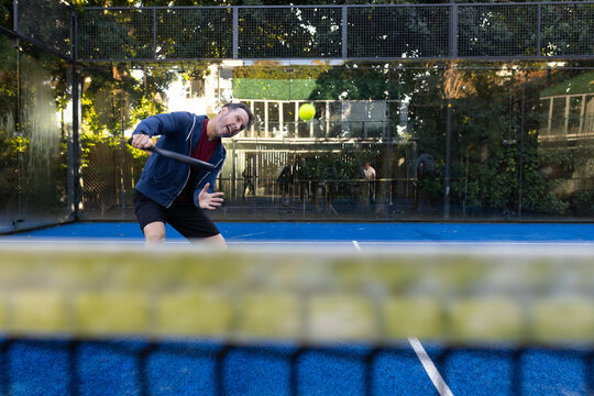 Adult man swinging paddle toward yellow ball on fenced blue court, blurred net, wearing navy hoodie