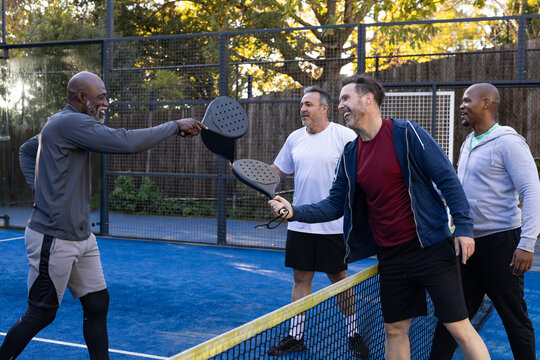 Diverse male players in sportswear tapping paddles and chatting at net on blue padel court