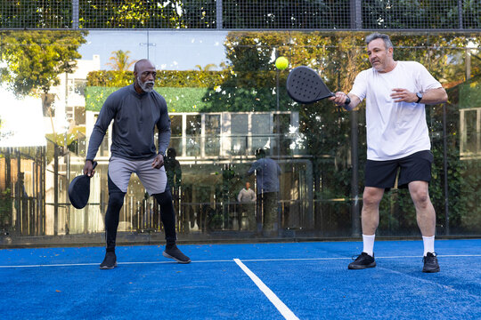 Diverse men in sportswear hitting yellow ball with padel rackets on blue court by glass wall