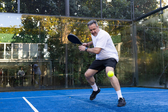 Middle-aged male lunging to strike yellow ball with black paddle on blue paddle court in sportswear