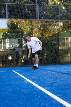 Sport-clad man bending and hitting yellow-ball with black-paddle on blue turf, glass reflecting