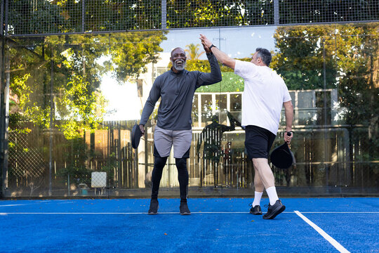 Senior and adult male friends high-fiving on blue paddle court with paddles, wearing leggings