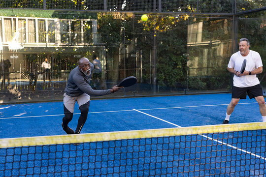 Diverse male pair lunging and returning yellow ball on blue court with black paddles