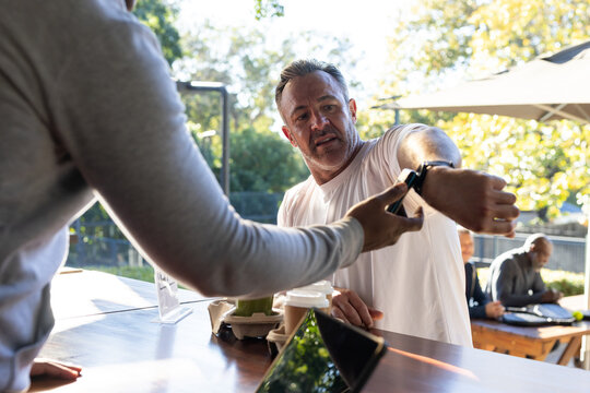 Diverse adults at cafe counter, customer extending left arm while staff scanning smartwatch