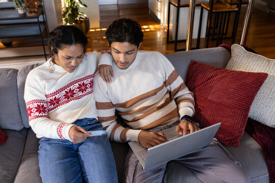African American couple sitting on gray sofa at home, woman holding receipt, man using laptop