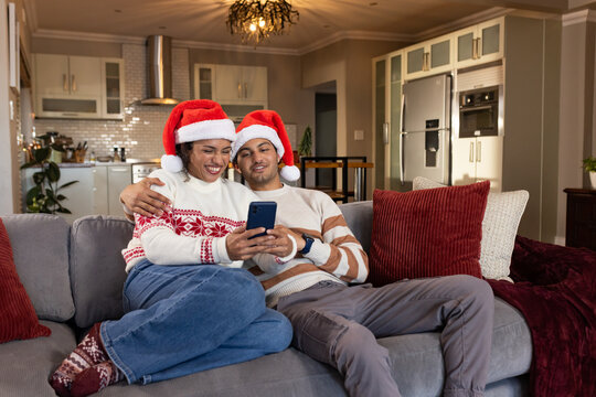 Couple sitting on gray sofa at home wearing Santa hats and festive sweaters, holding smartphone