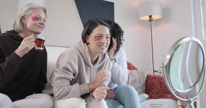 Diverse female friends sharing spa night, adjusting pink eye patches at mirror, sipping tea