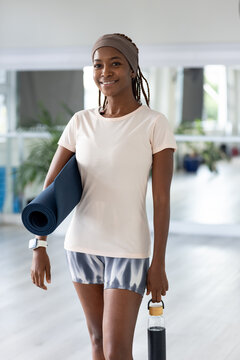 Female standing in yoga studio holding navy mat and water bottle, wearing headband, smartwatch