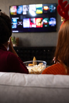 Two women sitting on couch watching wall-mounted TV, holding popcorn bowl, with red antler headband