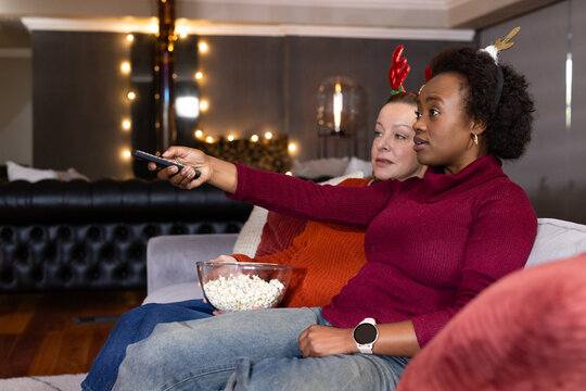Mature women friends on sofa wearing knit sweaters and antlers, sharing popcorn, pointing remote