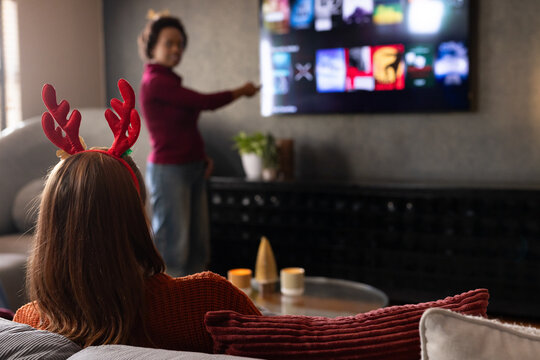Diverse female friends in festive sweaters browsing stream menu on wall TV with remote, copy space