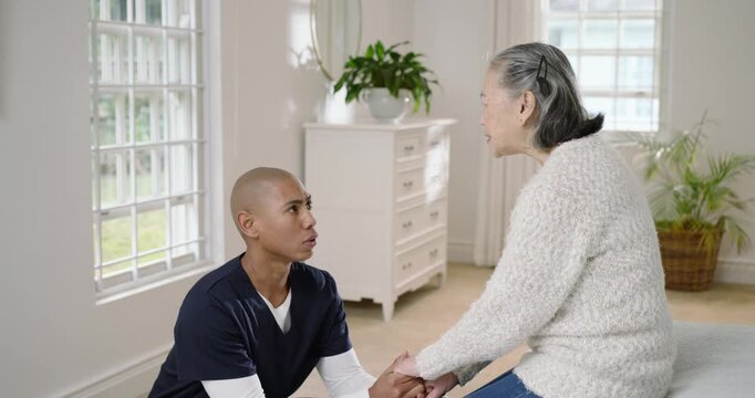 Caregiver kneeling, holding hands and speaking, helping Senior Chinese woman into standing by bed
