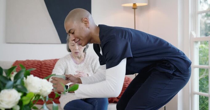 Senior Asian woman after knock, African American nurse entering, placing mug, helping sip on sofa