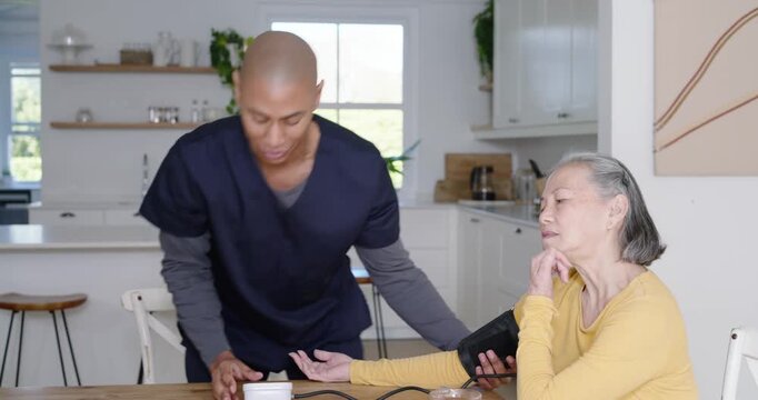African American nurse in scrubs coming to Senior Asian woman, fitting cuff at home, checking BP