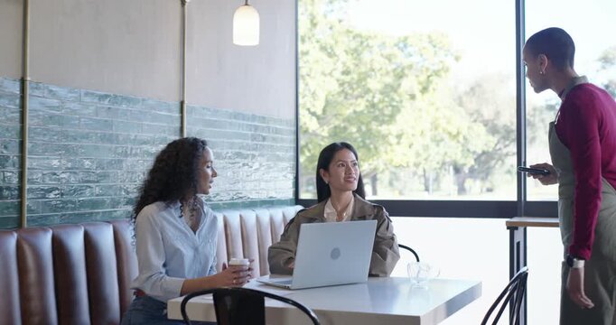 Diverse female coworkers tapping wrist to pay while server holding reader at cafe
