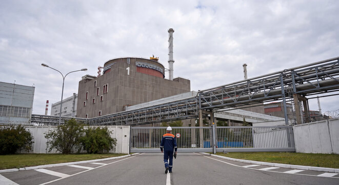Industrial technician walking along asphalt pathway beside massive pipeline infrastructure and nuclear power station buildings under overcast sky