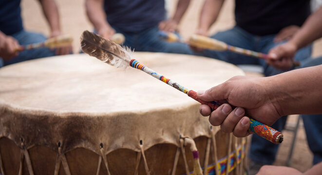 Close-up of several indigenous drummers striking a large traditional powwow drum with mallet drumsticks during a musical ritual ceremony outdoors.