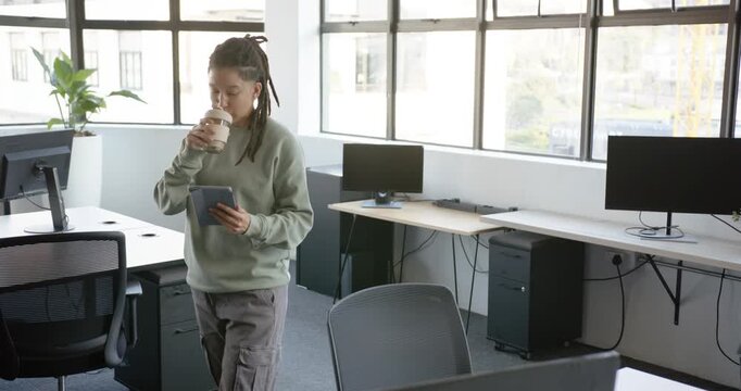 Nonbinary person entering from left into open office holding tablet sipping cup reading in sage top