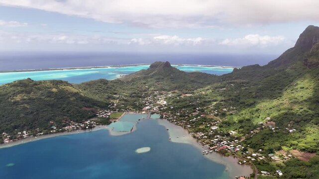 An aerial drone shot descends over the dramatic green peaks of Bora Bora, revealing a panoramic view of Vaitape village, its coastal harbor basins, and the distant blue ocean lagoon reef barrier