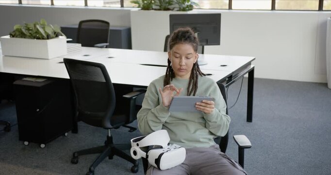 Non-binary sitting in open-plan office holding tablet, tapping to review work, VR headset on lap