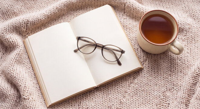 An open blank notebook rests with modern reading glasses and a hot cup of black tea on a soft beige knitted wool blanket creating a peaceful study scene