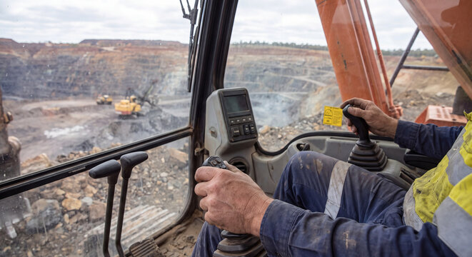 Interior view from the cabin of a heavy excavator with an operator handling joystick controls looking out over a vast open pit mining quarry on a sunny day