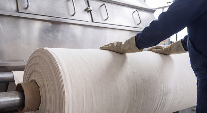Factory employee rolling large cylinder of white textile material on manufacturing production line inside modern industrial facility