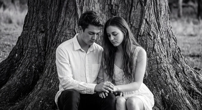 Monochrome portrait of an affectionate couple sitting closely together at the base of an ancient textured tree trunk in an outdoor natural setting.