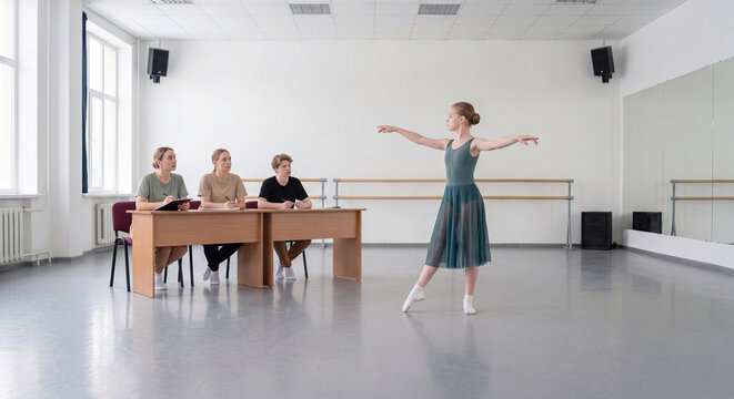 A graceful young ballerina performs an arabesque pose while three judges observe her routine from behind a table in a bright dance hall