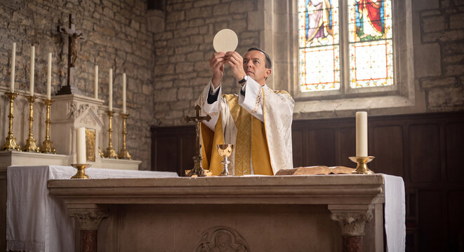 A priest raises the sacramental host above the altar candles during the consecration ritual of holy communion inside an ancient cathedral sanctuary