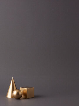 Minimalist still life arrangement of metallic gold geometric cone cube and sphere objects on a solid dark grey background with copy space