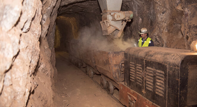 Mining cart locomotive driving into rocky underground mine shaft surrounded by stone tunnel walls and ambient dust clouds